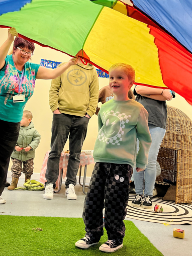 A young boy stands happily underneat a parachute held aloft by staff and parents.
