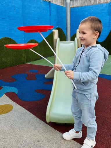 A young boy smiles shows off his magic skills, balancing two spinning red plates on sticks. 
