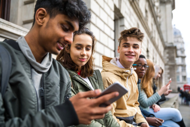 Three teenagers sat outside building with one looking at smartphone