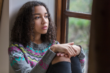 Teenage girl sat by window with her arms wrapped around her knees looking pensive