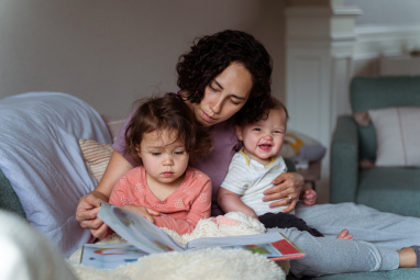 Mother reading with a child while also holding a baby who is smiling 