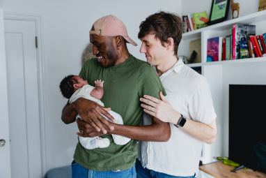 One man stands holding his baby while his partner holds the man's arm while looking at their baby