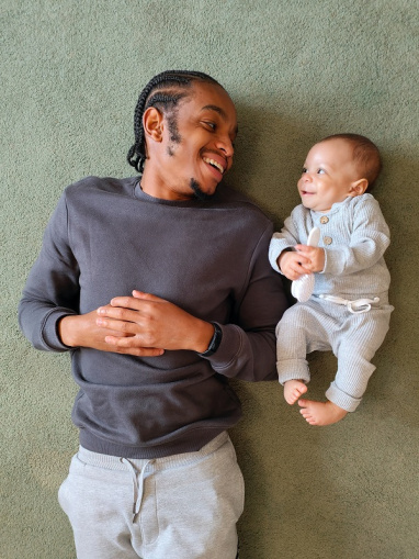 A father and his baby smile at each other while lying on a carpet.