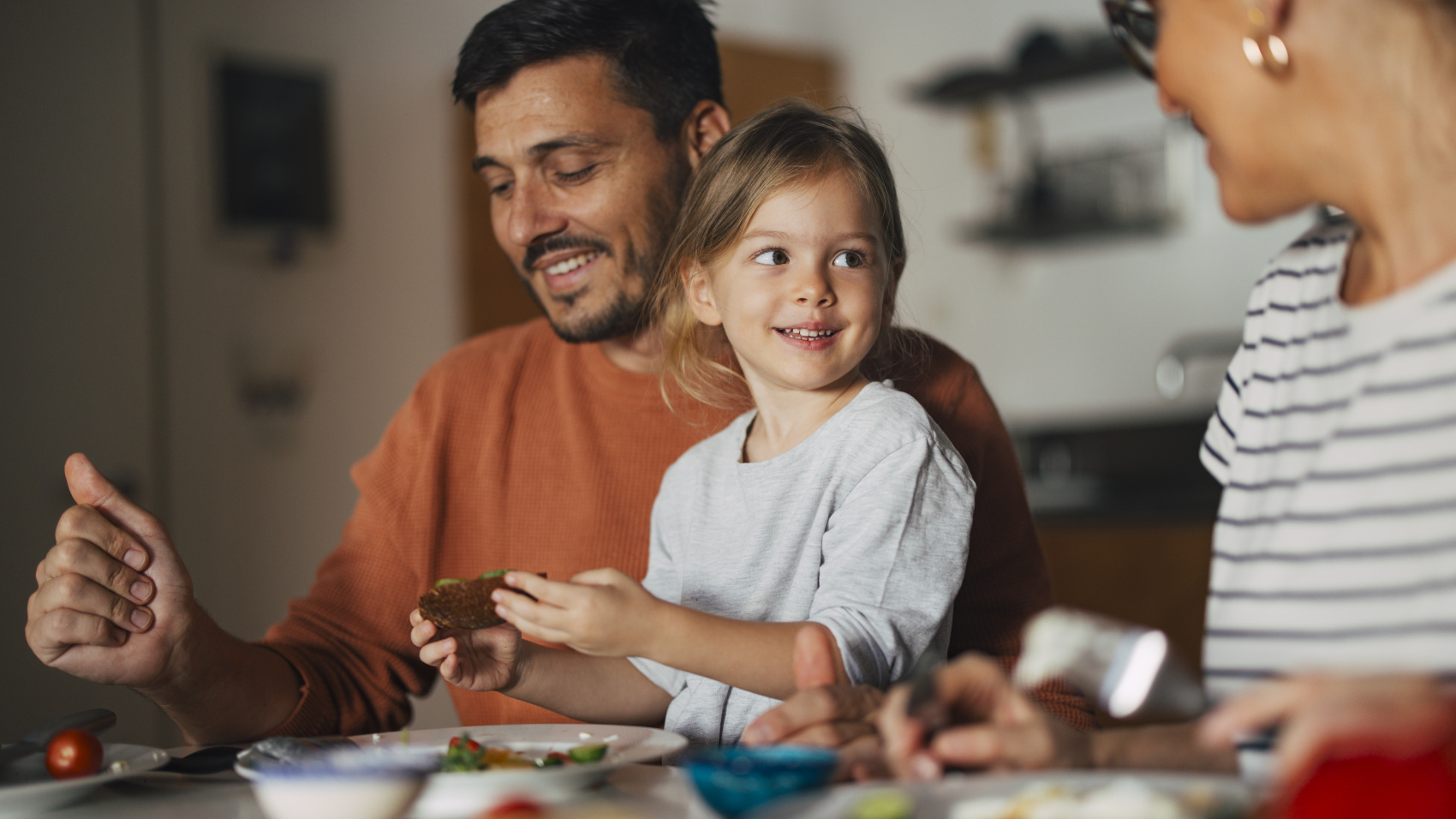 Man smiling with young girl on his lap at the table with woman smiling at them both 