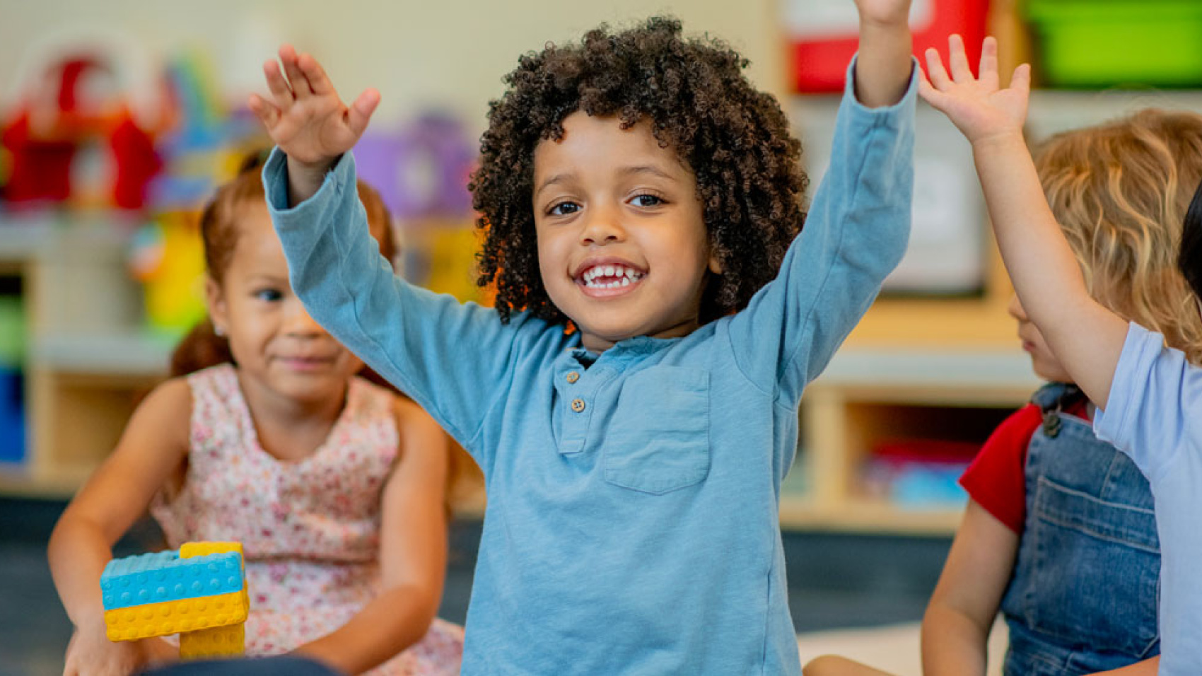 Toddler at play school raising arms and smiling joyfully at the camera while another child watches in the background