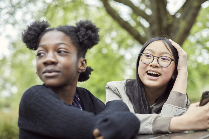 Two girls sit at a table outdoors and look to their right, smiling