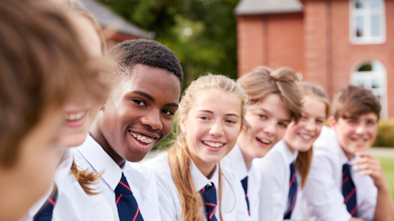 A group of pupils in school uniform, smile as they sit listening to one of their friends