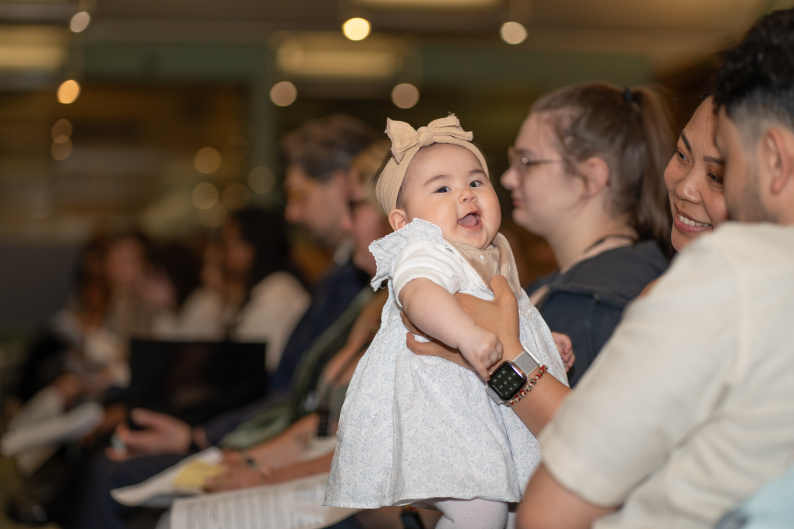 A baby held by her parents looks at the camera, at APPGC event