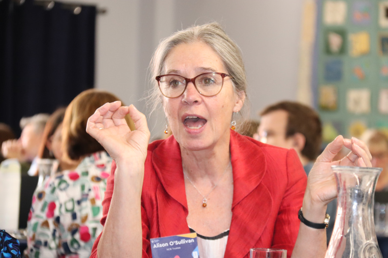 Woman with grey hair and glasses and a red jacket talking at a conference 