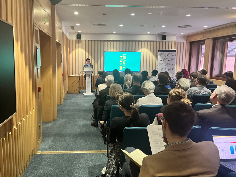 Professor Robbie Duschinsky stands at lectern and presents his research to the audience, who are sitting opposite him. A big screen reads 'British Library' showcasing the location.
