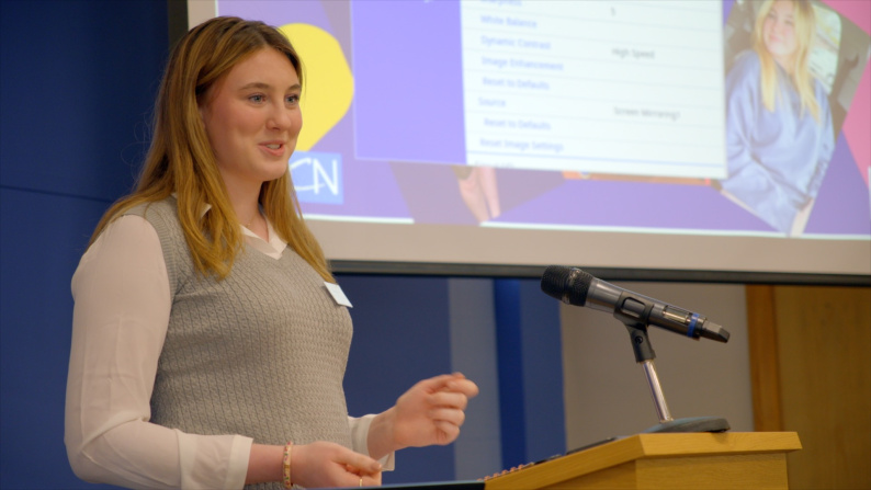 Woman presenting at a lectern in front of a screen 