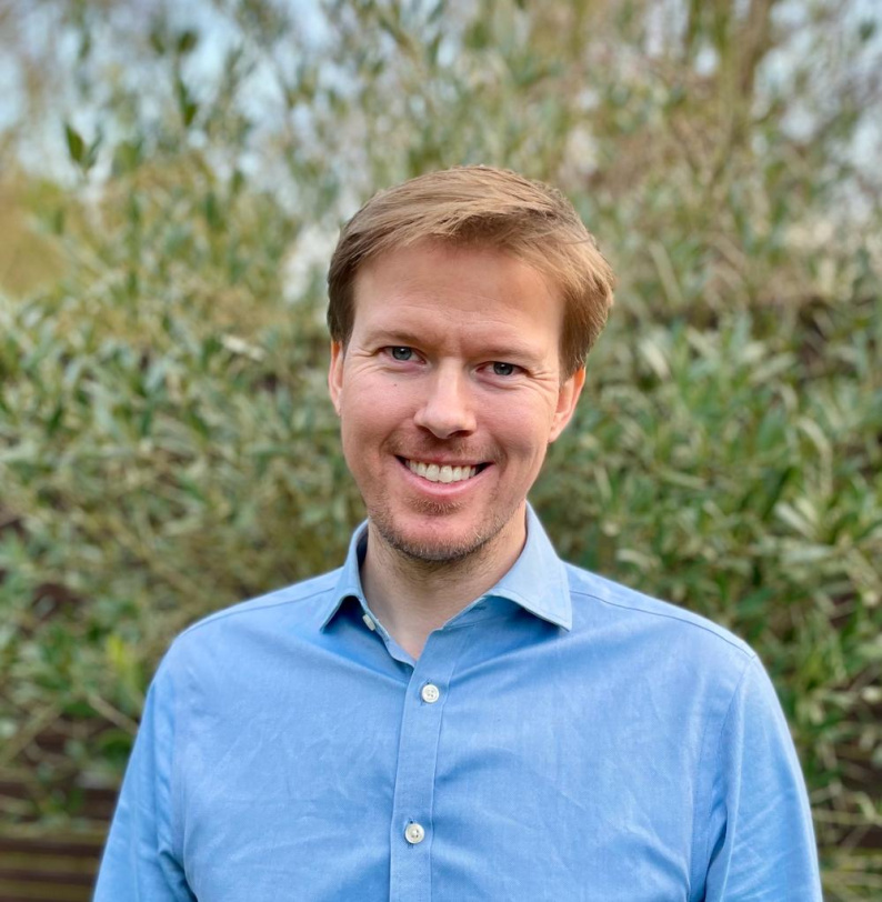 A picture of Phillip Anderson smiling wearing a blue shirt in front of green trees