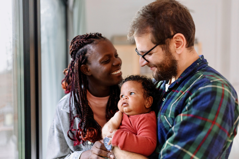 Man and a woman smiling and holding a baby in a home setting