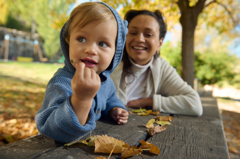 A young boy holding a leaf outside watched by a smiling adult