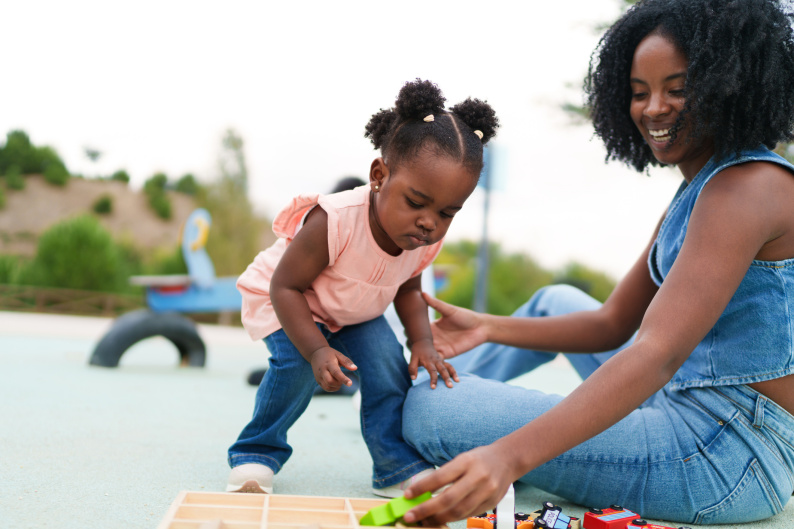 Early years age girl looking at toy held by smiling adult woman 